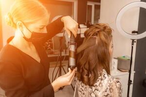 uma cabeleireiro dentro uma Preto mascarar prepara uma mulher, faz uma Penteado com cachos usando uma ondulação ferro. grandes luz Castanho natural cabelo. auto Cuidado conceito. foto