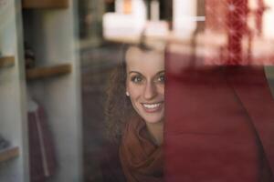 cativante sorrir do uma jovem mulher atrás uma cafeteria janela em uma brilhante dia foto