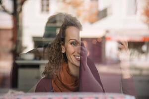 sorridente mulher com encaracolado cabelo desfrutando Visão a partir de atrás vidro janela em ensolarado dia foto