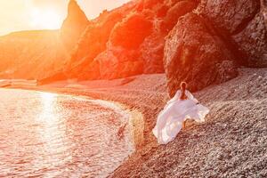 misterioso mulher silhueta grandes cabelo anda em em a de praia oceano água, mar ninfa vento escuta para a aceno. lança acima uma grandes branco vestir, uma divino pôr do sol. artístico foto a partir de a costas sem uma face