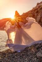 misterioso mulher silhueta grandes cabelo anda em em a de praia oceano água, mar ninfa vento escuta para a aceno. lança acima uma grandes branco vestir, uma divino pôr do sol. artístico foto a partir de a costas sem uma face