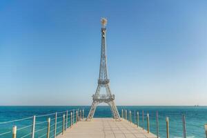 ampla modelo do a eiffel torre em a de praia. uma mulher anda em ao longo a cais para a torre, vestindo uma azul Jaqueta e branco jeans. foto