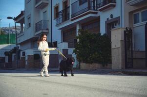 fofa criança menina caminhando dela cachorro em coleira. cheio comprimento retrato do uma feliz criança dentro branco roupa de esporte, desfrutando uma manhã andar com dela pedigree Preto de raça pura Cocker spaniel. eu sou levando meu animal para andar. foto