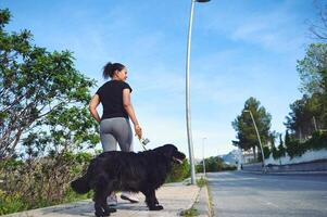 traseiro Visão do uma jovem ativo mulher caminhando dela cachorro em trela dentro a natureza em uma lindo ensolarado verão dia. bonita fêmea atleta corrida com dela animal dentro a manhã foto