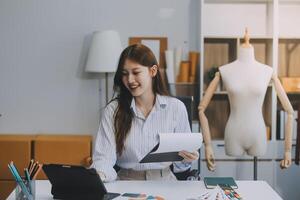 retrato lindo sorrir ásia desenhador mulher usar computador dentro alfaiate tecido moda pequeno o negócio conectados oficina. jovem proprietário comece empreendedor. criativo menina têxtil vestuário sme o negócio conceito foto