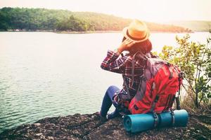 mulher jovem hippie com o mochileiro sentado na pedra, apreciando a bela vista da paisagem do lago e da montanha. este viajante felicidade com viagem em aventura de estilo de vida liberdade mulheres mais fortes foto