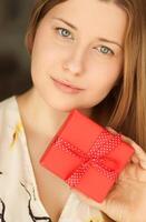 feliz sorridente mulher segurando uma vermelho presente caixa, face retrato com natural Maquiagem e feriado estilo de vida às casa foto