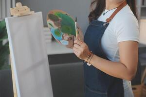 cortada imagem do fêmea artista em pé dentro frente do a cavalete e mergulho escova para dentro cor paleta foto