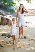 jovem mulher mãe com uma pequeno filha dentro branco vestidos tendo uma Diversão em Beira Mar dentro a sombra do árvores e Palmeiras. Alto qualidade foto