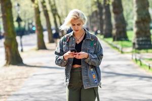 jovem urbana com penteado moderno usando smartphone, andando na rua em um parque urbano. foto