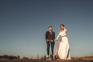 casal de noivos sorridente andando de scooters ao longo da estrada foto