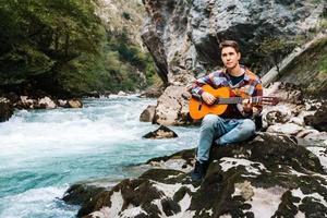 jovem tocando violão sentado na margem de um rio de montanha em um fundo de rochas e floresta foto