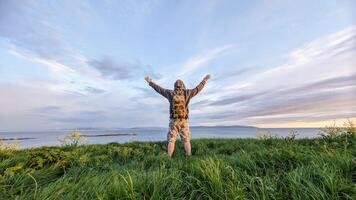 homem caminhante dentro camo calção e mochila em topo do verde colina, mãos acima, Visão em selvagem atlântico caminho às galway, Irlanda, liberdade, aventura e estilo de vida conceito, natureza fundo foto