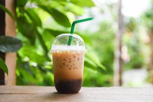 Café com leite gelado em copo de plástico na mesa de madeira e fundo verde da natureza - bebidas geladas foto