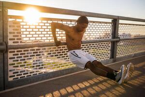 jovem afro-americano homem é exercício em a ponte dentro a cidade. ele é fazendo marcha ré flexões. foto