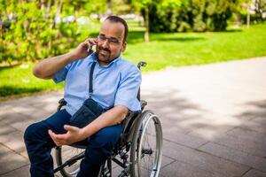 retrato do feliz homem dentro cadeira de rodas. ele é desfrutando ensolarado dia dentro cidade parque e falando em telefone. foto