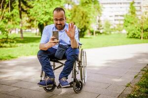 retrato do feliz homem dentro cadeira de rodas. ele é desfrutando ensolarado dia dentro cidade parque e Mensagens em Smartphone. foto