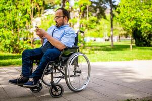 retrato do homem dentro cadeira de rodas. ele é desfrutando ensolarado dia dentro cidade parque e Mensagens em Smartphone. foto