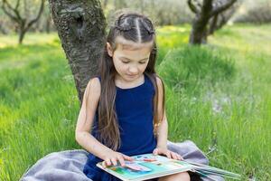 uma criança menina é lendo uma livro dentro a parque foto