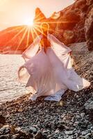 misterioso mulher silhueta grandes cabelo anda em em a de praia oceano água, mar ninfa vento escuta para a aceno. lança acima uma grandes branco vestir, uma divino pôr do sol. artístico foto a partir de a costas sem uma face