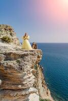 mulher amarelo vestir mar. lado Visão jovem lindo sensual mulher dentro amarelo grandes vestir posando em uma Rocha Alto acima a mar às pôr do sol. menina dentro natureza contra a azul céu foto