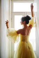 uma mulher silhueta dentro uma dourado luxuoso vestir contra a fundo do uma janela detém uma cortina com dela mãos. elegante senhora dentro uma amarelo grandes seda vestir com nu voltar, costas visualizar. foto