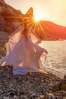 misterioso mulher silhueta grandes cabelo anda em em a de praia oceano água, mar ninfa vento escuta para a aceno. lança acima uma grandes branco vestir, uma divino pôr do sol. artístico foto a partir de a costas sem uma face