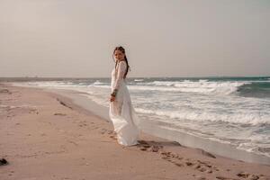 modelo dentro boho estilo dentro uma branco grandes vestir e prata joalheria em a de praia. dela cabelo é trançado, e lá estão muitos pulseiras em dela braços. foto