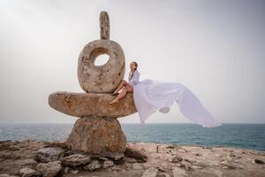 uma mulher senta em uma pedra escultura fez do ampla pedras. ela é vestido dentro uma branco grandes vestir, contra a pano de fundo do a mar e céu. a vestir desenvolve dentro a vento. foto