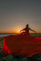 uma mulher dentro uma vermelho vestir é em pé dentro uma campo com a Sol configuração atrás dela. ela é alcançando acima com dela braços estendido, Como E se ela é tentando para pegar a Sol. a cena é sereno e pacífico. foto