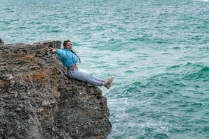 uma mulher dentro uma azul Jaqueta senta em uma Rocha acima uma penhasco acima a mar, olhando às a tormentoso oceano. menina viajante descansa, acha, sonhos, goza natureza. Paz e calma paisagem, ventoso clima. foto