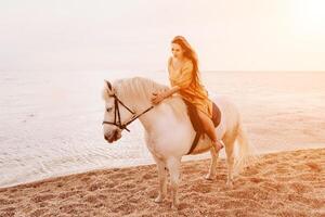 uma mulher dentro uma vestir carrinhos Próximo para uma branco cavalo em uma praia, com a azul céu e mar dentro a fundo. foto