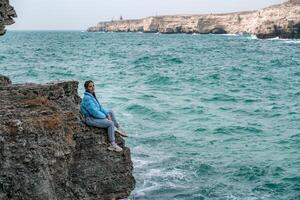 uma mulher dentro uma azul Jaqueta senta em uma Rocha acima uma penhasco acima a mar, olhando às a tormentoso oceano. menina viajante descansa, acha, sonhos, goza natureza. Paz e calma paisagem, ventoso clima. foto