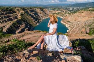 uma mulher dentro uma branco vestir é sentado em uma Rocha negligenciar uma corpo do água. a cena é sereno e pacífico, com a mulher desfrutando a Visão e a calma do a arredores. foto