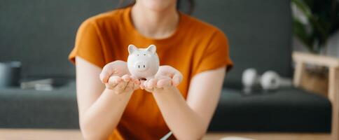 ásia mulheres mão salvando uma moeda para dentro porquinho banco com de madeira para cálculo e tábua em a mesa para negócios, finança, salvando dinheiro e propriedade investimento conceito. foto