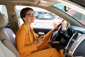 à moda morena mulher com curto corte de cabelo dirigindo uma carro, segurando uma copo do café. vestindo amarelo Jaqueta e óculos. foto