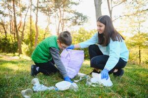 mulher voluntário e pequeno Garoto colheita acima a plástico lixo e colocando isto dentro biodegradável saco do lixo ao ar livre. ecologia, reciclando e proteção do natureza conceito. de Meio Ambiente proteção. foto