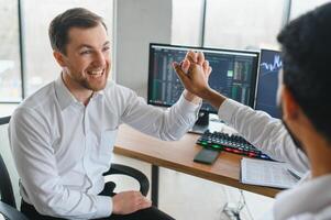 dois homens comerciantes sentado às escrivaninha às escritório juntos monitoramento ações dados vela gráficos em tela analisando preço fluxo sorridente alegre tendo lucro trabalho em equipe conceito foto