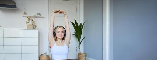 vertical tiro do sorridente, ativo e saudável mulher exercite-se às lar, segue ginástica instruções, relógios em computador portátil dentro fones de ouvido, alongar dela braços foto