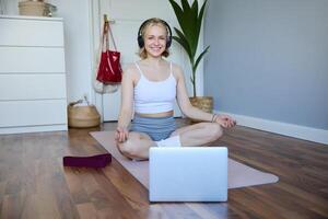 retrato do em forma e saudável mulher às lar, prática ioga, sentado em borracha esteira, ouvindo para instruções on-line, usando meditação música para relaxar, Segue orientação em computador portátil foto