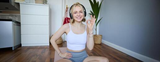 retrato do fofa sorridente loiro mulher vestindo sem fio fones de ouvido, exercite-se às casa em borracha ioga esteira, sentado dentro uma sala, mostrando OK, sim mão placa foto