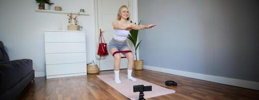retrato do jovem Atlético mulher, vlogger fazer uma sobre casa dar certo, ginástica instrutor tiroteio quão para Faz perna exercícios, usando elástico resistência banda, usando ioga esteira foto