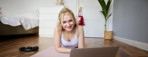 retrato do lindo loiro mulher olhando às ginástica tutoriais em computador portátil, deitado em borracha ioga esteira, Segue exercite-se instruções conectados foto