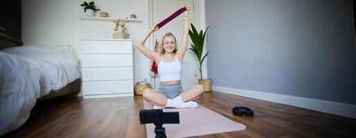 sorridente jovem loiro mulher gravação sobre dar certo, tiroteio em digital Câmera, ginástica instrutor mostra quão para usar resistência banda durante exercícios foto