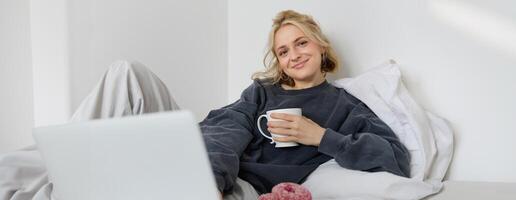 retrato do feliz mulher desfrutando folga às lar, deitado dentro cama, assistindo televisão mostrar em computador portátil, olhando às tela e sorridente, bebendo chá e comendo dentro quarto foto