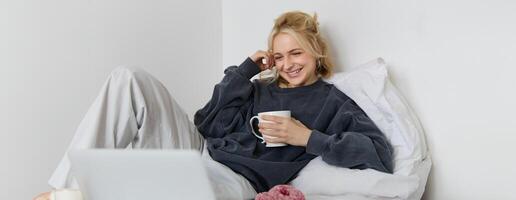 retrato do sincero, feliz jovem mulher deitado dentro cama, olhando às computador portátil tela, segurando copo do chá e comendo rosquinha, ficando às casa em fim de semana, gastos bastante Tempo sozinho foto