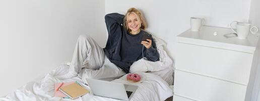 retrato do jovem mulher, aluna estudando dentro dela cama, relaxante enquanto preparando trabalho de casa, comendo rosquinha, usando computador portátil dentro quarto e bebendo chá foto