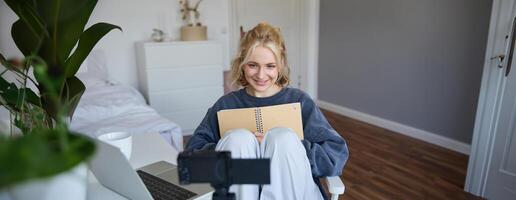 retrato do sorridente fofa mulher, estilo de vida blogueiro, senta dentro dela quarto com diariamente Diário ou planejador, registros em digital Câmera, cria conteúdo para social meios de comunicação sobre diariamente rotina foto