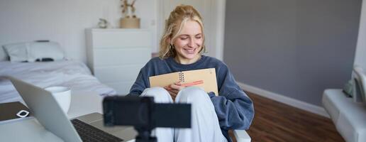 retrato do sorridente fofa mulher, estilo de vida blogueiro, senta dentro dela quarto com diariamente Diário ou planejador, registros em digital Câmera, cria conteúdo para social meios de comunicação sobre diariamente rotina foto