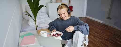 retrato do sorridente jovem loiro mulher dentro fones de ouvido, sentado dentro sala, assistindo filme em computador portátil, comendo café da manhã e bebendo chá, tendo almoço dentro frente do computador tela foto